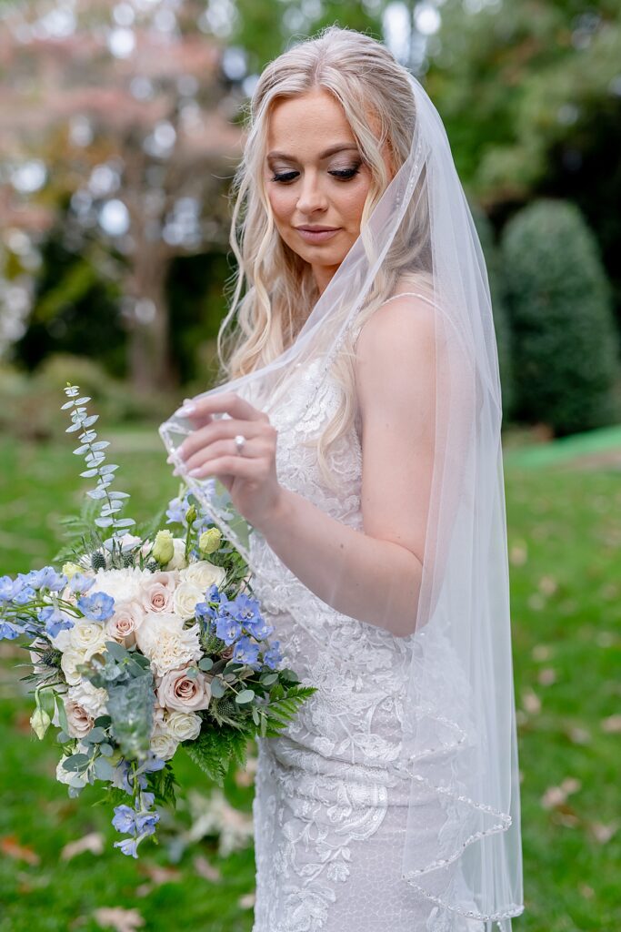 Bride touches tip of her veil while holding her bouquet of light blue, pastel pink roses, and cream flowers.