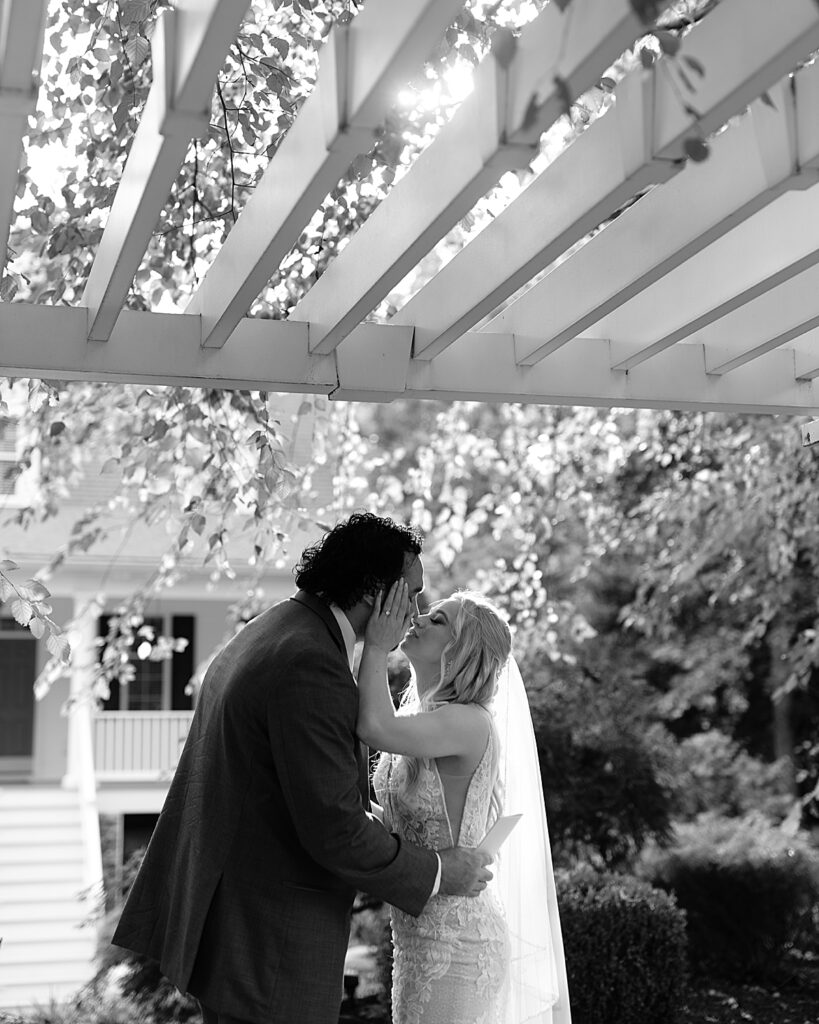 Bride and groom kiss under the sunlit pergola in a garden at their venue, captured by Maryland wedding photographer Nadine Nasby Photography