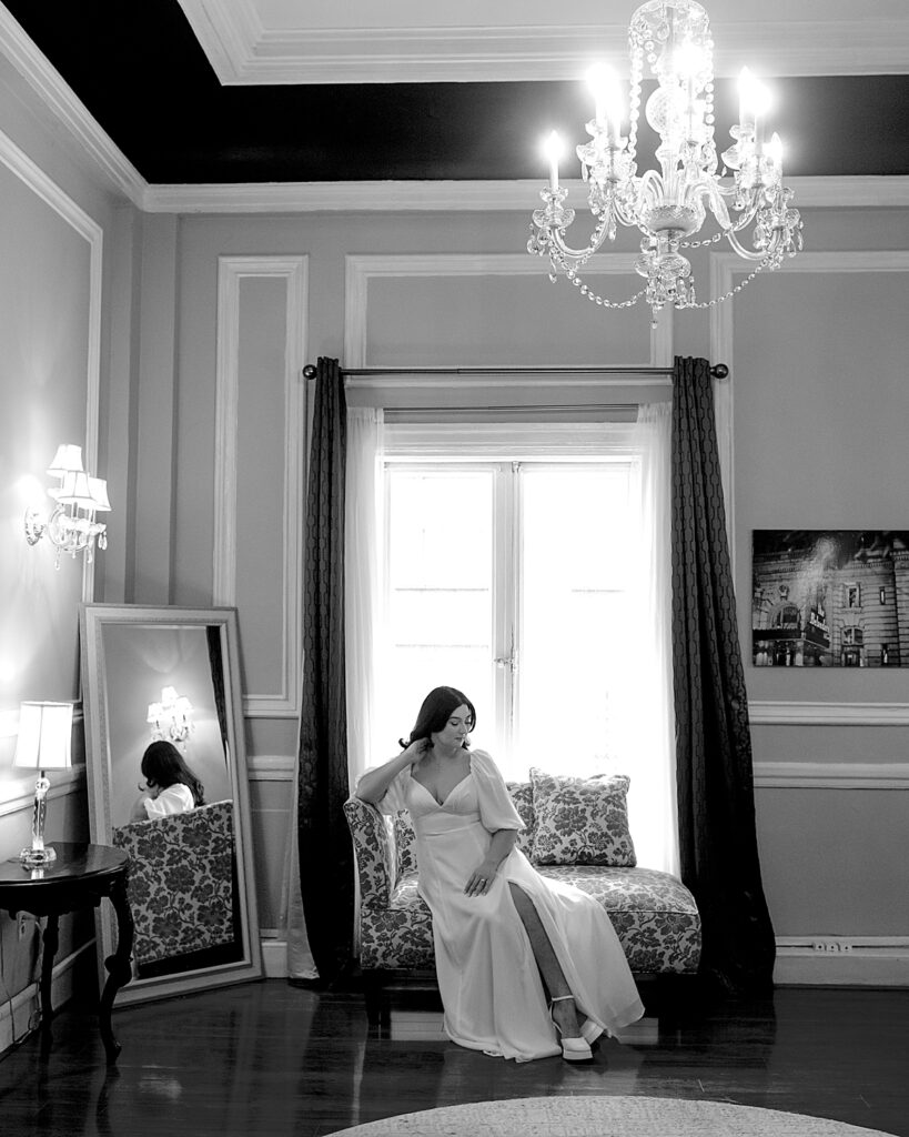 Bride sits on settee in front of window with mirrored reflected behind her and antique chandelier hanging above - documentary style wedding photographer DC
