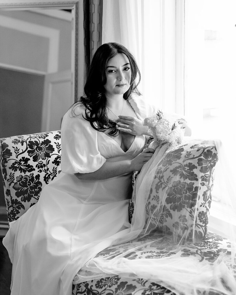 Bride holds necklace while she sits on settee by the window - documentary style wedding photographer DC